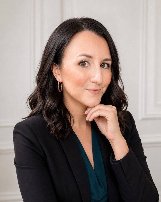 Studio portrait of Janie Windmiller, founder of Be The Good Marketing, in a black blazer over a deep green top, resting her chin on her hand in front of a white paneled wall